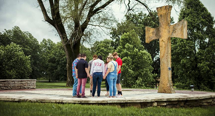 A group of five people stands in a circle holding hands near a large stone cross outdoors, surrounded by trees and greenery.