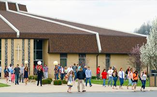 A group of people, including men, 女性, and children, exit a modern church building with brown roof and cross symbol in an outdoor setting. Trees and shrubs decorate the surroundings.